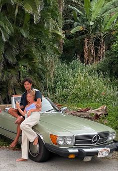 two people sitting on the hood of a car with a woman standing next to it