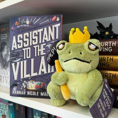 a stuffed frog with a crown sitting on top of a bookshelf next to other books