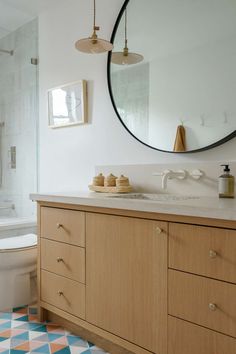 a white toilet sitting next to a bathroom sink under a round mirror on top of a wooden cabinet