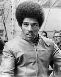 a black and white photo of a man with an afro wearing a leather jacket in front of other people