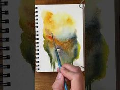 a person holding a pencil in their left hand and painting on paper with watercolors