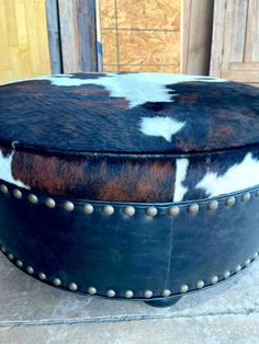 a cow hide ottoman with rivets and studded trim on the top, sitting on concrete