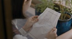 a woman holding a piece of paper with writing on it next to a potted plant