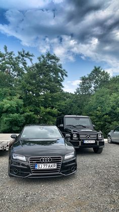 two black cars parked next to each other in front of some trees and sky with clouds