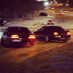 two black cars driving down a street next to snow covered ground and buildings at night