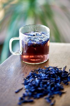 a glass cup filled with blue tea next to a pile of dried lavender on top of a wooden table