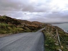 an empty road on the side of a hill with water in the distance and clouds overhead