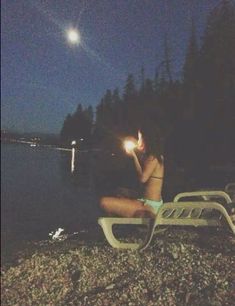 a woman sitting on a lawn chair in front of the water at night with her hand up to her face