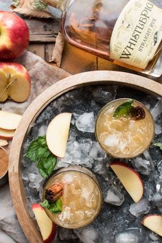 two glasses filled with ice and apple slices on top of an old fashioned wooden tray