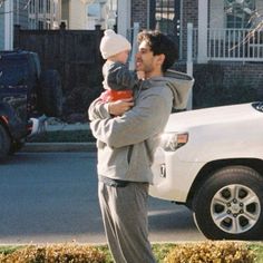 a man holding a baby in his arms while standing next to a white car on the street