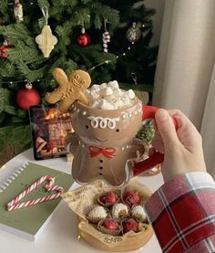 a person holding up a cup filled with marshmallows next to a christmas tree