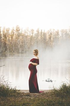 a pregnant woman standing in front of a body of water wearing a long red dress