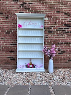 a white shelf with flowers on it next to a brick wall and vase filled with pink flowers