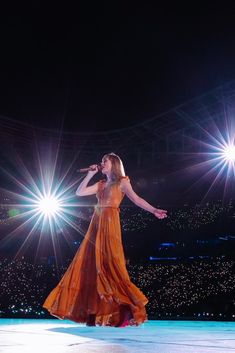 a woman in an orange dress on stage with lights behind her and one hand out