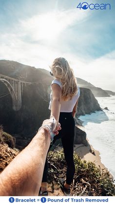 a man and woman holding hands while standing on top of a cliff overlooking the ocean