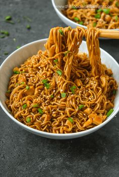 someone holding chopsticks over noodles in a white bowl with green onions on the side