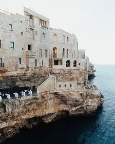 people are standing on the edge of a cliff by the water in front of some buildings