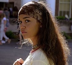 a woman with long hair wearing a bandana and looking off into the distance while standing in front of a building