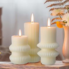 three white candles sitting on top of a table next to vases filled with flowers