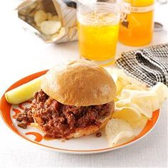 a sloppy joe sandwich on a plate with potato chips and orange juice in the background