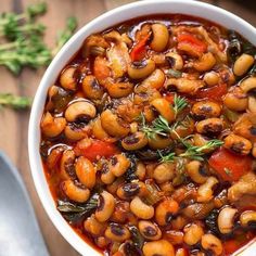 a white bowl filled with beans and vegetables on top of a wooden table next to a spoon