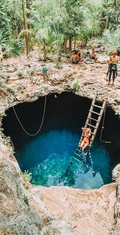 two people climbing up a ladder into a deep blue hole in the ground surrounded by trees