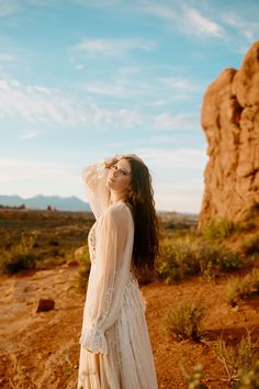 a woman standing in the desert wearing a long white dress and holding her hair back