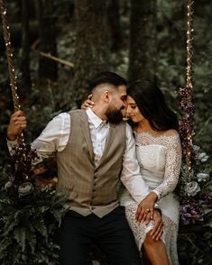 a man and woman sitting on a swing in the woods, holding each other's hands