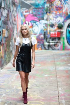 a woman walking down a street with graffiti on the walls behind her and wearing a black leather dress