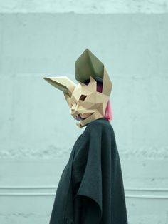 a person wearing a paper mask and cape with pink hair standing in front of a wall