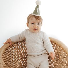 a baby sitting in a wicker basket wearing a birthday hat