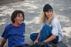 two young men sitting on skateboards in the street smiling at each other's faces