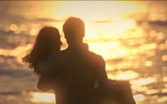 a man and woman standing on the beach with their arms around each other as the sun sets