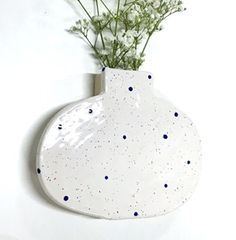 a white vase with blue dots and flowers in it hanging on the side of a wall