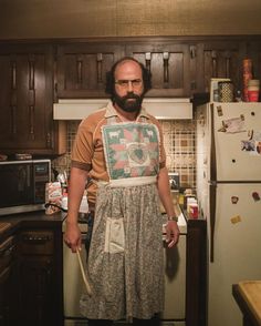 a bearded man in an apron stands in the kitchen with his hands on his hips