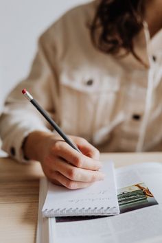 a woman sitting at a table with a book and pen in her hand while writing