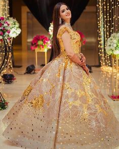 a woman in a gold and white gown standing on a stage with flowers behind her