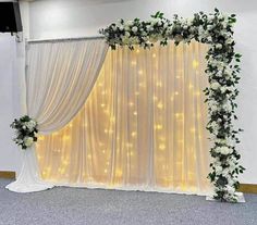 a white backdrop with flowers and greenery on the side is decorated with lights for a wedding ceremony