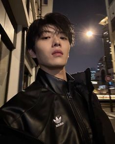 a young man wearing a black leather jacket standing in front of a building at night