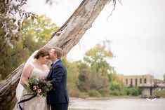 a bride and groom standing next to each other in front of a tree by the water