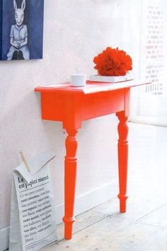 an orange table with flowers on it in front of a white wall and a newspaper