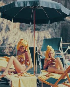 two women in bathing suits sitting on beach chairs under an umbrella and looking at their cell phones