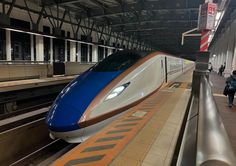 a blue and white train pulling into a station with people walking on the platform next to it