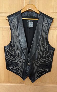 Wonderful black North Beach Leather Vest by Michael Hoban with Western Cowboy style stitching, snap and back belt buckle.  The leather is very soft and pliable, and there's slight padding behind the stitching in front.  There are 2 pockets on the outside of the vest and 2 more on the inside lining, as shown in the photos.  The only flaw I can find is a slight rough spot and 2 tiny pin dots on the right lapel.  They look more prominent in the photos than in real life.  Otherwise, the vest is in excellent condition.  A size 42, the measurements are approx:   16 1/2' across the shoulders, 42" across the chest   40" across the bottom    25" long in front to the vest tips   22" long in the mid back See my entire shop collection here https://www.etsy.com/shop/RobinClaytonVintage Cowboy Leather Vest, Gothic Cowboy Fashion, Men’s Vests, Goth Cowboy Men, Unique Outfits Men, Black Cowboy Outfit, Western Gothic Fashion, Edgy Western Style, Goth Vest