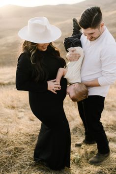 a man and woman holding a baby in their arms while standing on top of a dry grass field