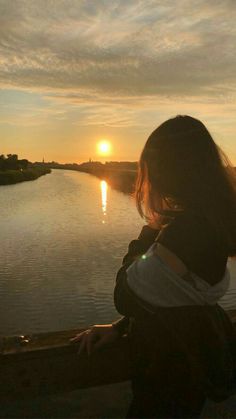 a woman sitting on the edge of a bridge watching the sun go down over water