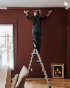 a woman is standing on a ladder and painting the wall in her living room with red walls