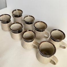 six coffee mugs are lined up on a white counter top, one is black and the other has brown speckles