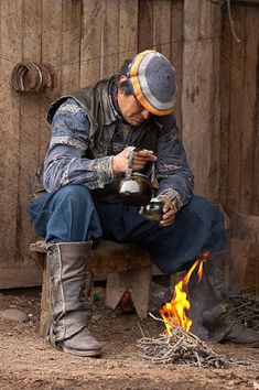 a man sitting on a bench next to a fire with a cup in his hand