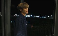 a young man standing in front of a window at night with the city lights behind him